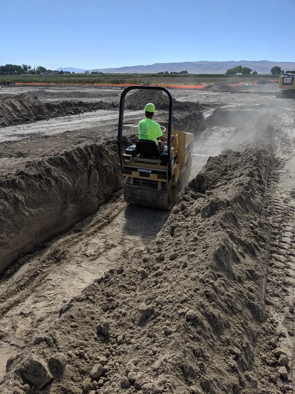 A Caterpillar smooth-drum compactor running a sub-grade pass in an open trench, operator in high-vis, Southern Idaho fields and mountains beyond