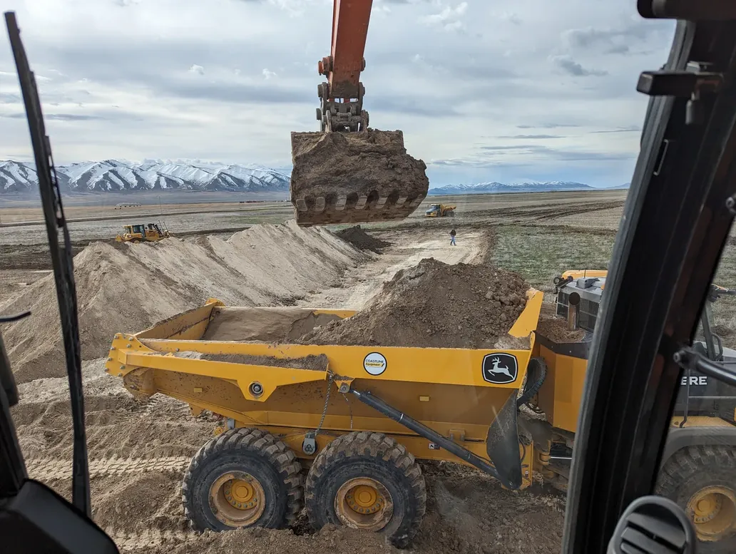 An excavator loading an articulated dump truck with cut earth on a Southern Idaho site, snow-capped mountains in the distance