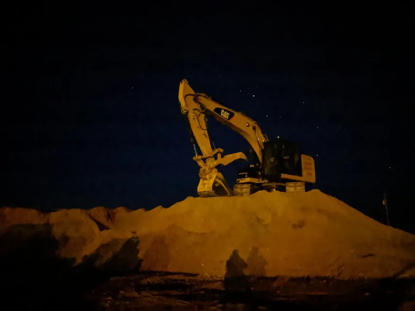 A Cat excavator silhouetted against a starry night sky on top of a dirt pile, with two crew members in the foreground, Dimond Excavation running long after dark